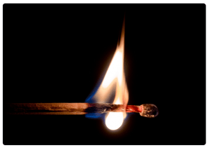A close-up of a matchstick igniting with a bright flame against a dark background.